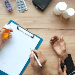 Vista cenital de un farmaceutico en su escritorio sosteniendo un frasco de pastillas de color naranja y tomando notas en un portapapeles blanco, mientras atiende y toma la mano de un paciente mayor. En la mesa de madera se observan un estetoscopio, un oxímetro de pulso, blísteres de cápsulas y frascos de medicina blancos, elementos clave en la atención personalizada para fidelizar clientes en la farmacia.