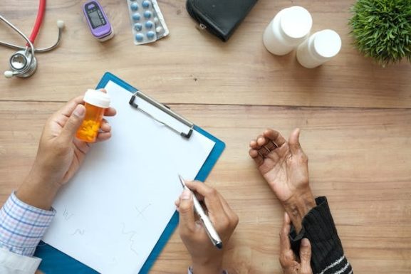Vista cenital de un farmaceutico en su escritorio sosteniendo un frasco de pastillas de color naranja y tomando notas en un portapapeles blanco, mientras atiende y toma la mano de un paciente mayor. En la mesa de madera se observan un estetoscopio, un oxímetro de pulso, blísteres de cápsulas y frascos de medicina blancos, elementos clave en la atención personalizada para fidelizar clientes en la farmacia.