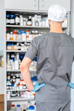 Vista desde atrás de un farmacéutico o profesional de la salud con uniforme gris, gorro blanco y guantes azules, de pie con las manos en las caderas mientras observa una estantería repleta de frascos y envases de medicamentos organizados, mostrando el abastecimiento necesario para fidelizar clientes en la farmacia mediante un servicio eficiente.