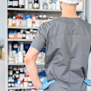 Vista desde atrás de un farmacéutico o profesional de la salud con uniforme gris, gorro blanco y guantes azules, de pie con las manos en las caderas mientras observa una estantería repleta de frascos y envases de medicamentos organizados, mostrando el abastecimiento necesario para fidelizar clientes en la farmacia mediante un servicio eficiente.