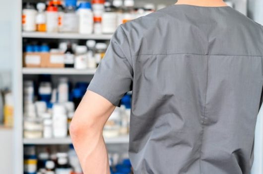 Vista desde atrás de un farmacéutico o profesional de la salud con uniforme gris, gorro blanco y guantes azules, de pie con las manos en las caderas mientras observa una estantería repleta de frascos y envases de medicamentos organizados, mostrando el abastecimiento necesario para fidelizar clientes en la farmacia mediante un servicio eficiente.