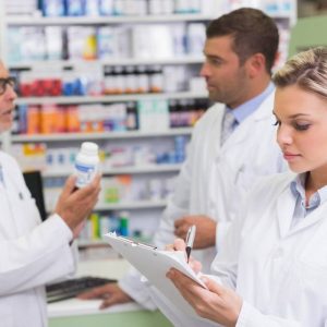 Tres profesionales de la salud con batas blancas trabajan en el interior de una botica; en primer plano, una farmacéutica rubia toma notas con un bolígrafo en un portapapeles, mientras al fondo dos compañeros conversan y revisan detalladamente un frasco de medicina blanco frente a las estanterías de productos, ejemplificando la coordinación interna y los canales de comunicación en una farmacia.