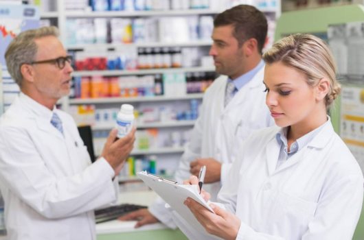 Tres profesionales de la salud con batas blancas trabajan en el interior de una botica; en primer plano, una farmacéutica rubia toma notas con un bolígrafo en un portapapeles, mientras al fondo dos compañeros conversan y revisan detalladamente un frasco de medicina blanco frente a las estanterías de productos, ejemplificando la coordinación interna y los canales de comunicación en una farmacia.