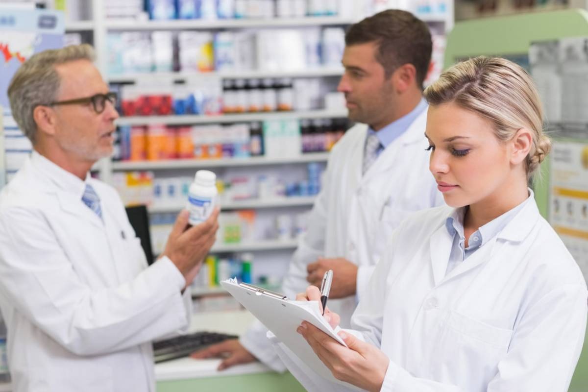 Tres profesionales de la salud con batas blancas trabajan en el interior de una botica; en primer plano, una farmacéutica rubia toma notas con un bolígrafo en un portapapeles, mientras al fondo dos compañeros conversan y revisan detalladamente un frasco de medicina blanco frente a las estanterías de productos, ejemplificando la coordinación interna y los canales de comunicación en una farmacia.