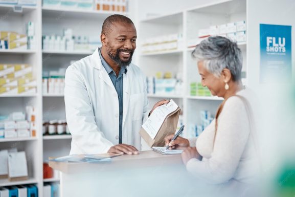 Un farmacéutico sonriente con bata médica blanca y barba atiende en el mostrador a una clienta de cabello corto y gris, sosteniendo una bolsa de papel kraft con una receta grapada. La mujer firma un documento en un portapapeles con un bolígrafo, ilustrando la interacción directa y la gestión del servicio de entrega como parte de los canales de comunicación en una farmacia. Al fondo se observan estanterías blancas con medicamentos y un cartel azul que dice "FLU SHOTS".