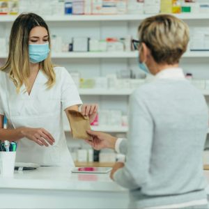 Una farmacéutica con uniforme blanco y mascarilla médica quirúrgica azul entrega una pequeña bolsa de papel kraft a una clienta de espaldas en el mostrador de atención. Al lado se observa un monitor de computadora y de fondo estanterías blancas repletas de productos de salud, mostrando la atención segura y el protocolo de técnicas de comunicación con el cliente en la farmacia.