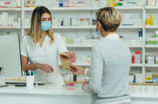 Una farmacéutica con uniforme blanco y mascarilla médica quirúrgica azul entrega una pequeña bolsa de papel kraft a una clienta de espaldas en el mostrador de atención. Al lado se observa un monitor de computadora y de fondo estanterías blancas repletas de productos de salud, mostrando la atención segura y el protocolo de técnicas de comunicación con el cliente en la farmacia.