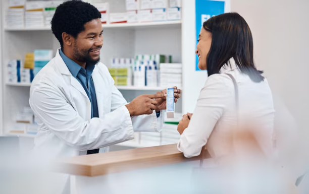 Un farmacéutico sonriente con bata blanca señala detalladamente las indicaciones en el empaque de una medicina azul y blanca mientras atiende a una mujer en el mostrador. Esta interacción personalizada en la botica, con estanterías de medicamentos de fondo, ejemplifica el uso de buenas técnicas de comunicación con el cliente en la farmacia.