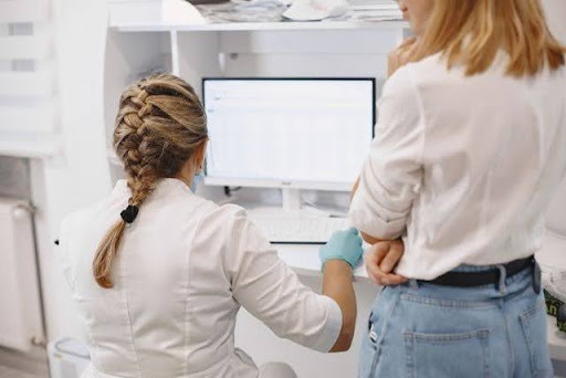 Vista desde atrás de una profesional farmacéutica con bata blanca, cabello trenzado y guantes azules sentada frente a un ordenador utilizando un crm farmacia, mientras una clienta de pie con camisa blanca y jeans observa la pantalla.