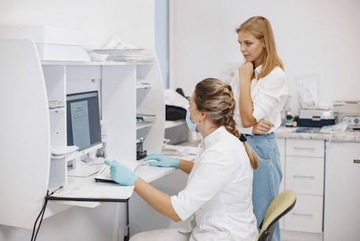 Vista lateral de una farmacéutica con bata blanca, mascarilla y guantes azules ingresando datos en un crm farmacia usando el teclado de la computadora en un mostrador blanco, junto a una mujer rubia que mira atentamente el monitor.